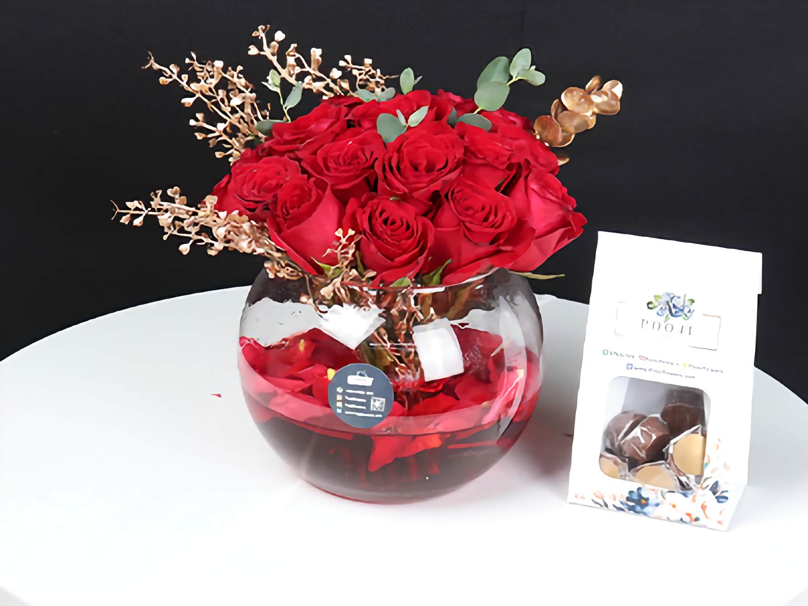 Bouquet of red roses in a glass vase with gold accents on a white surface, next to a small box of chocolates.