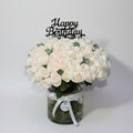 Bouquet of white roses in a glass vase with a 'Happy Birthday' candle on a wooden table.