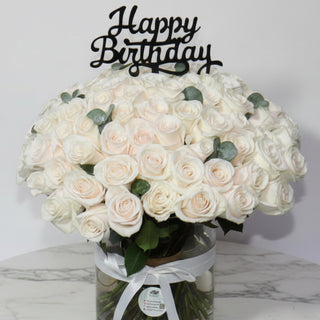 Bouquet of white roses in a glass vase with a 'Happy Birthday' candle on a wooden table.