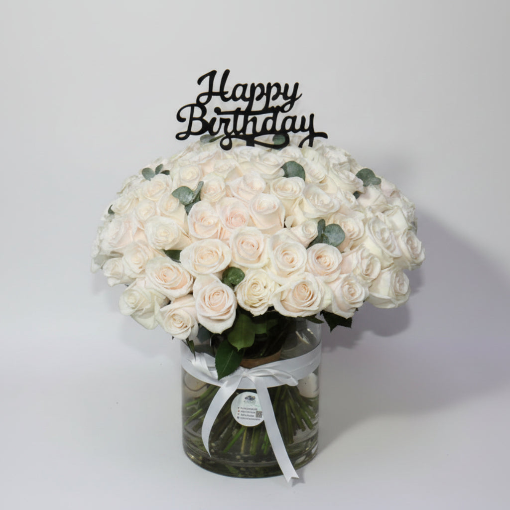 Bouquet of white roses in a glass vase with a 'Happy Birthday' candle on a wooden table.