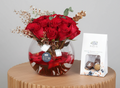 Bouquet of red roses in a glass vase with gold accents on a white surface, next to a small box of chocolates.