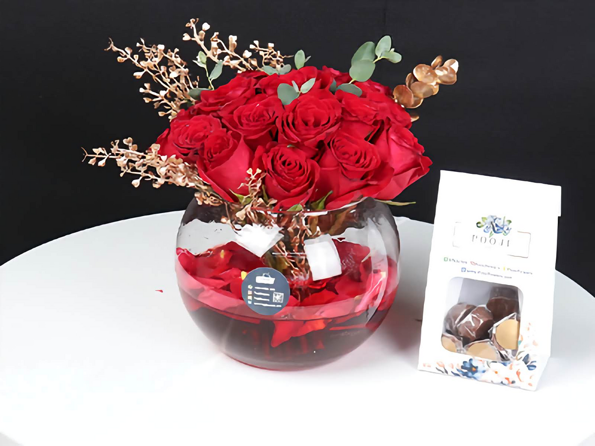 Bouquet of red roses in a glass vase with gold accents on a white surface, next to a small box of chocolates.