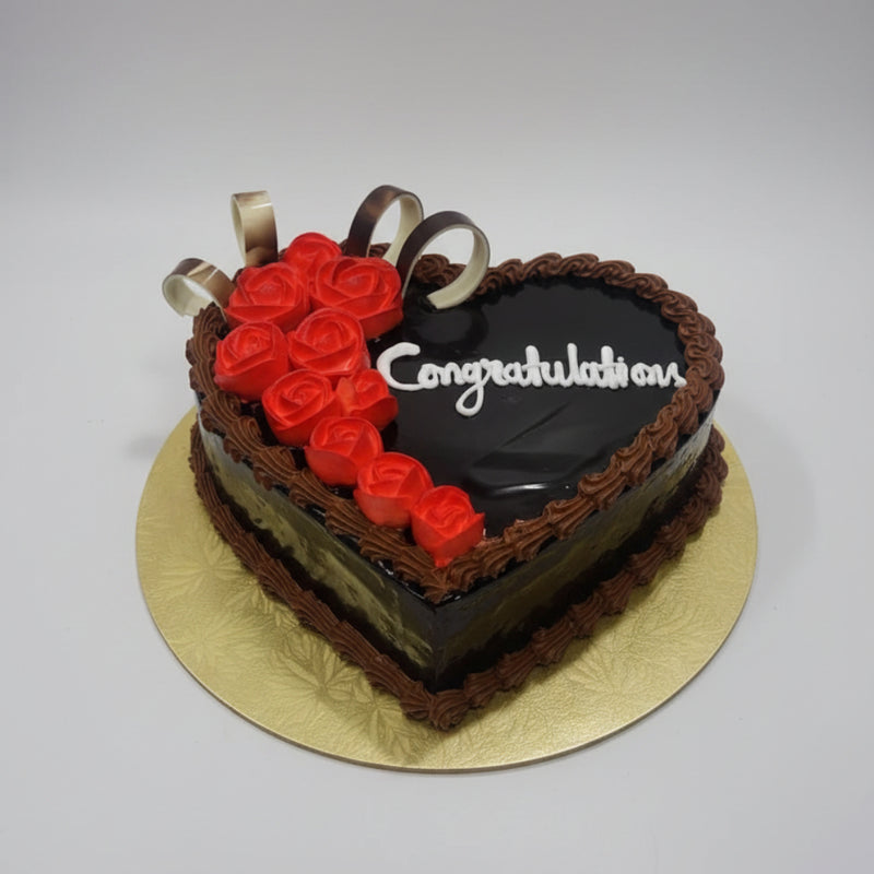 A heart-shaped chocolate cake with red rose decorations and white 'Congratulations' icing, displayed on a gold cake board on a stainless steel countertop.