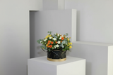 Floral arrangement in a black container on a white pedestal against a gray wall.