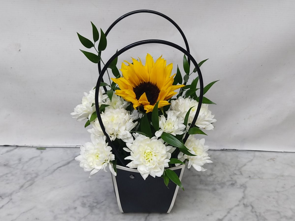 Floral arrangement with a sunflower and white flowers in a black basket on a marble surface.