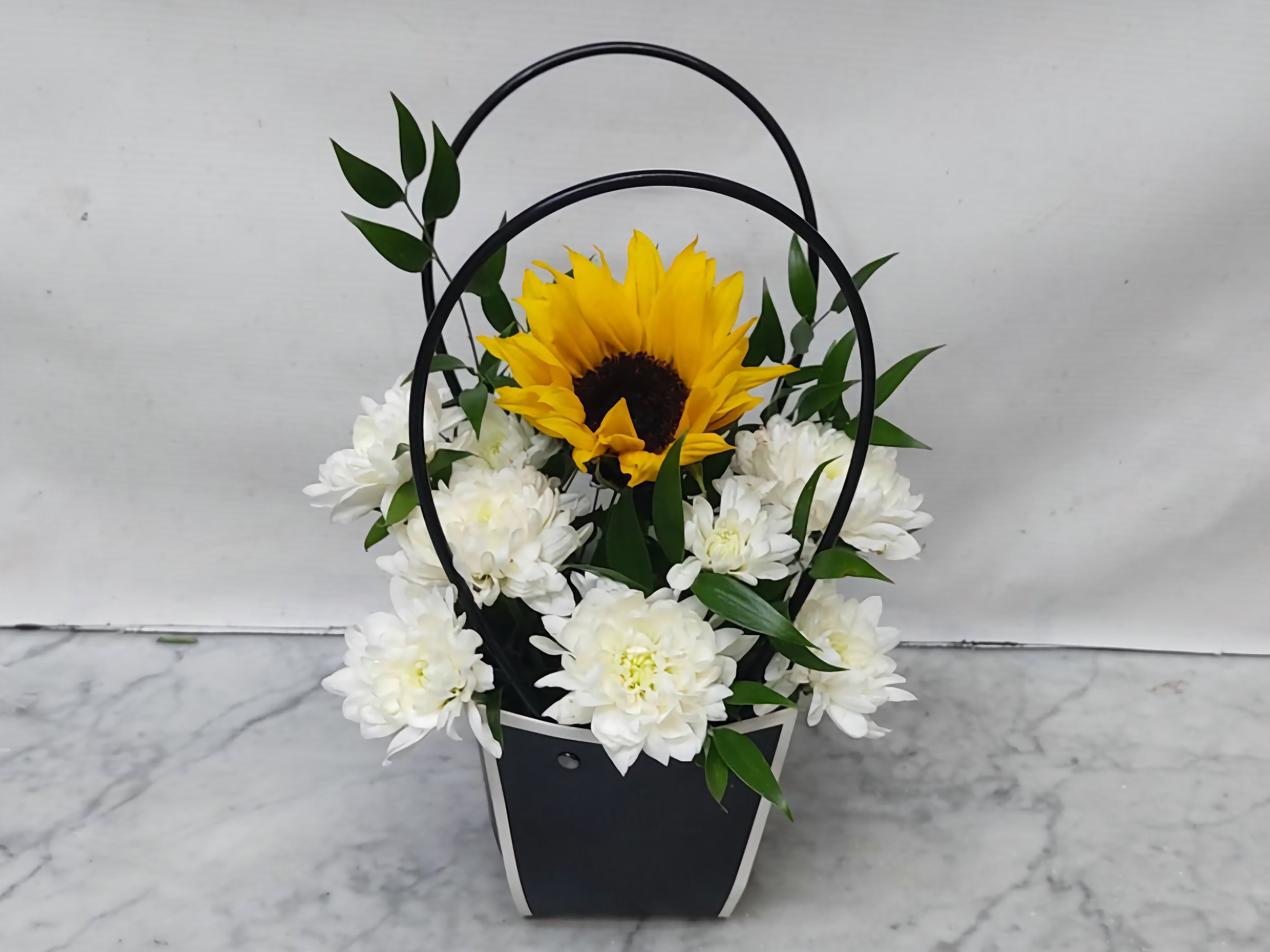 Floral arrangement with a sunflower and white flowers in a black basket on a marble surface.