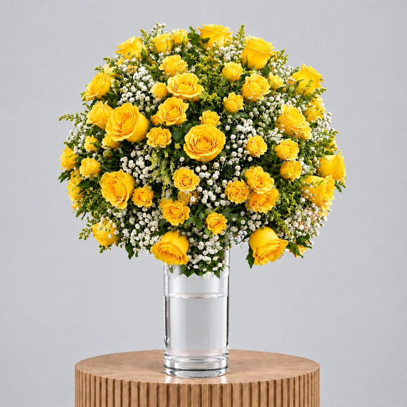 A round, lush bouquet of yellow roses and white baby's breath arranged in a clear glass vase, displayed on a round wooden pedestal against a solid gray background.