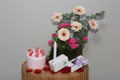 Small pink cake with cookies, a candle, and a bouquet of flowers on a wooden table against a gray background.