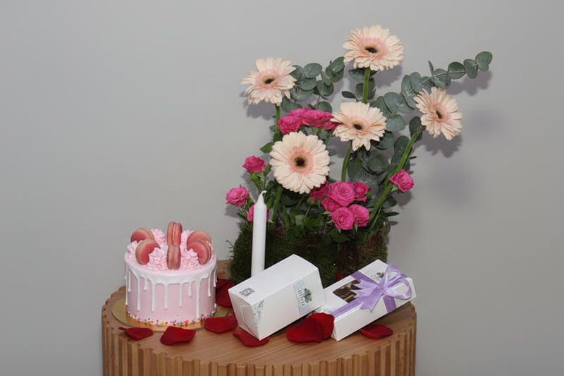 Small pink cake with cookies, a candle, and a bouquet of flowers on a wooden table against a gray background.