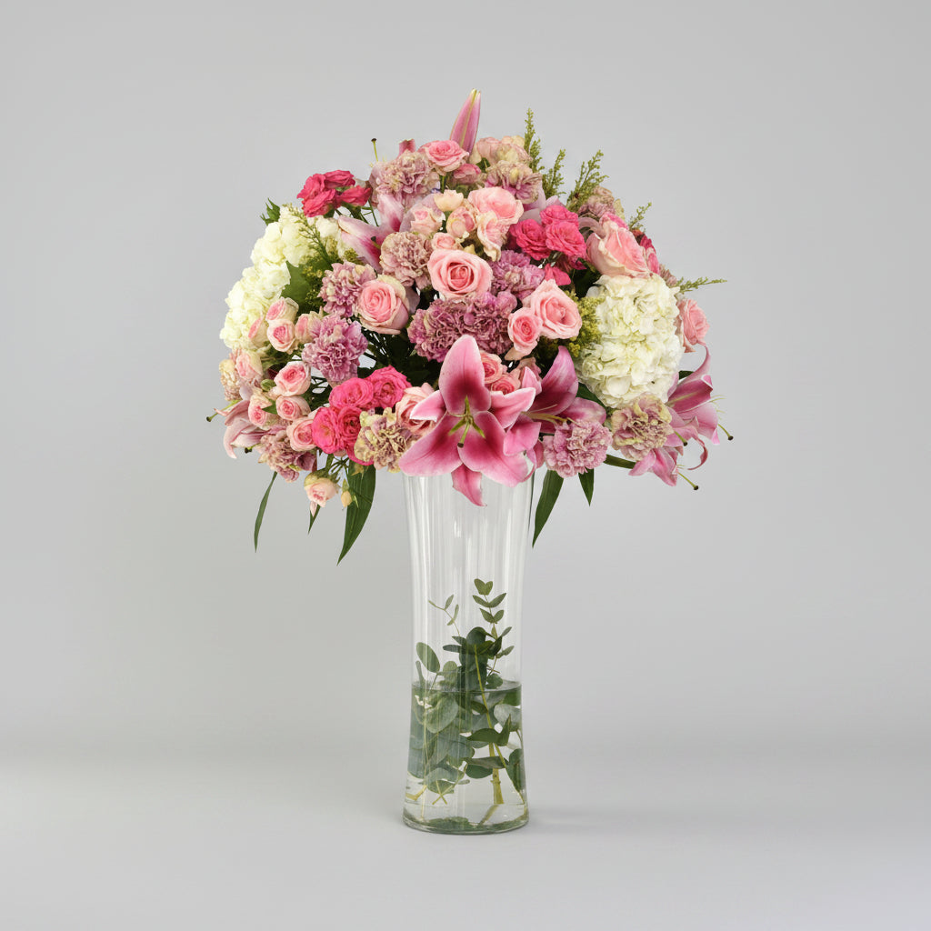 A vibrant floral arrangement featuring pink and white flowers in a tall, clear glass vase, set on a light marble surface against a white wall with soft window shadows.