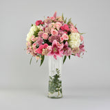 A vibrant floral arrangement featuring pink and white flowers in a tall, clear glass vase, set on a light marble surface against a white wall with soft window shadows.
