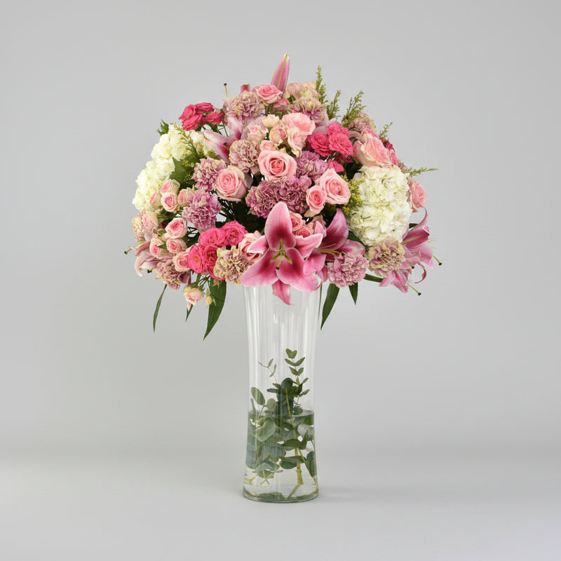 A vibrant floral arrangement featuring pink and white flowers in a tall, clear glass vase, set on a light marble surface against a white wall with soft window shadows.