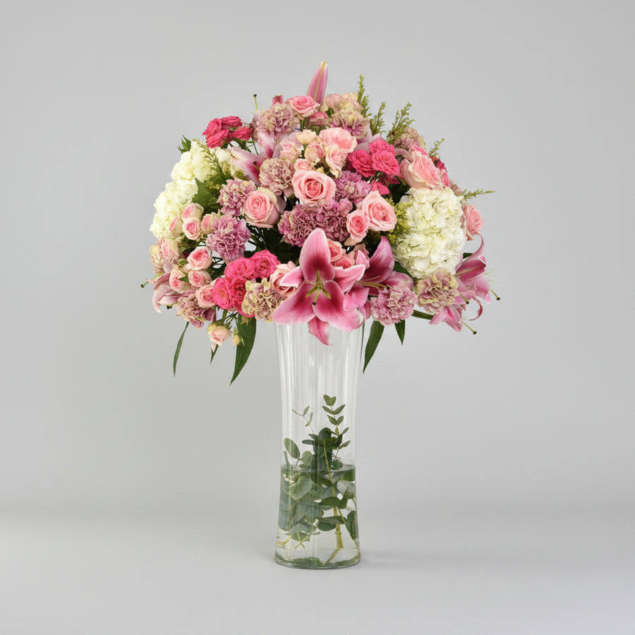 A vibrant floral arrangement featuring pink and white flowers in a tall, clear glass vase, set on a light marble surface against a white wall with soft window shadows.