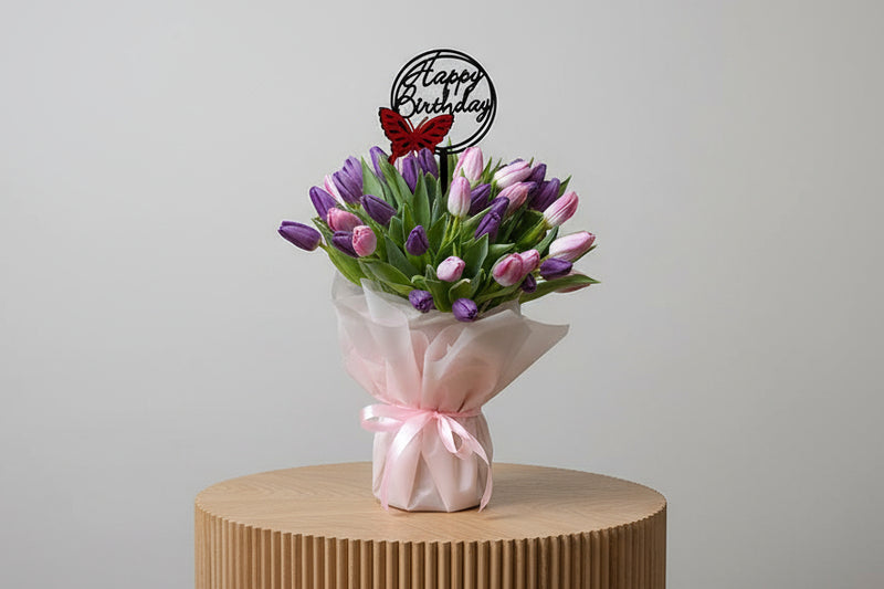 Bouquet of purple and pink tulips with a 'Happy Birthday' candle on a wooden stand.
