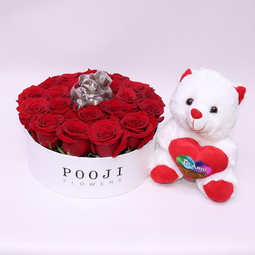Red roses in a box with a white teddy bear wearing a heart-shaped 'Te Amo' pillow on a light gray background.