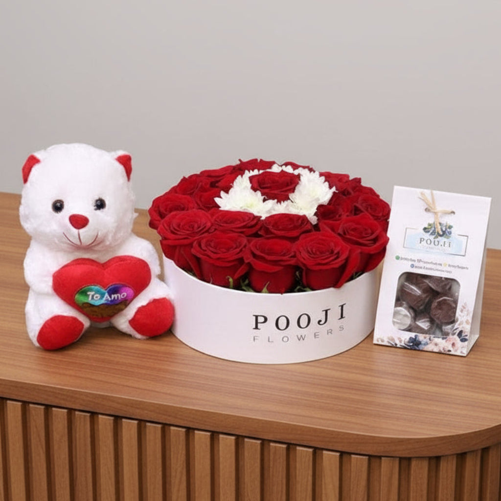 White teddy bear with a red heart, box of red roses, and a card on a wooden surface.