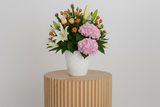 Bouquet of flowers with pink hydrangeas, white lilies, and orange roses in a white vase on a light gray background.