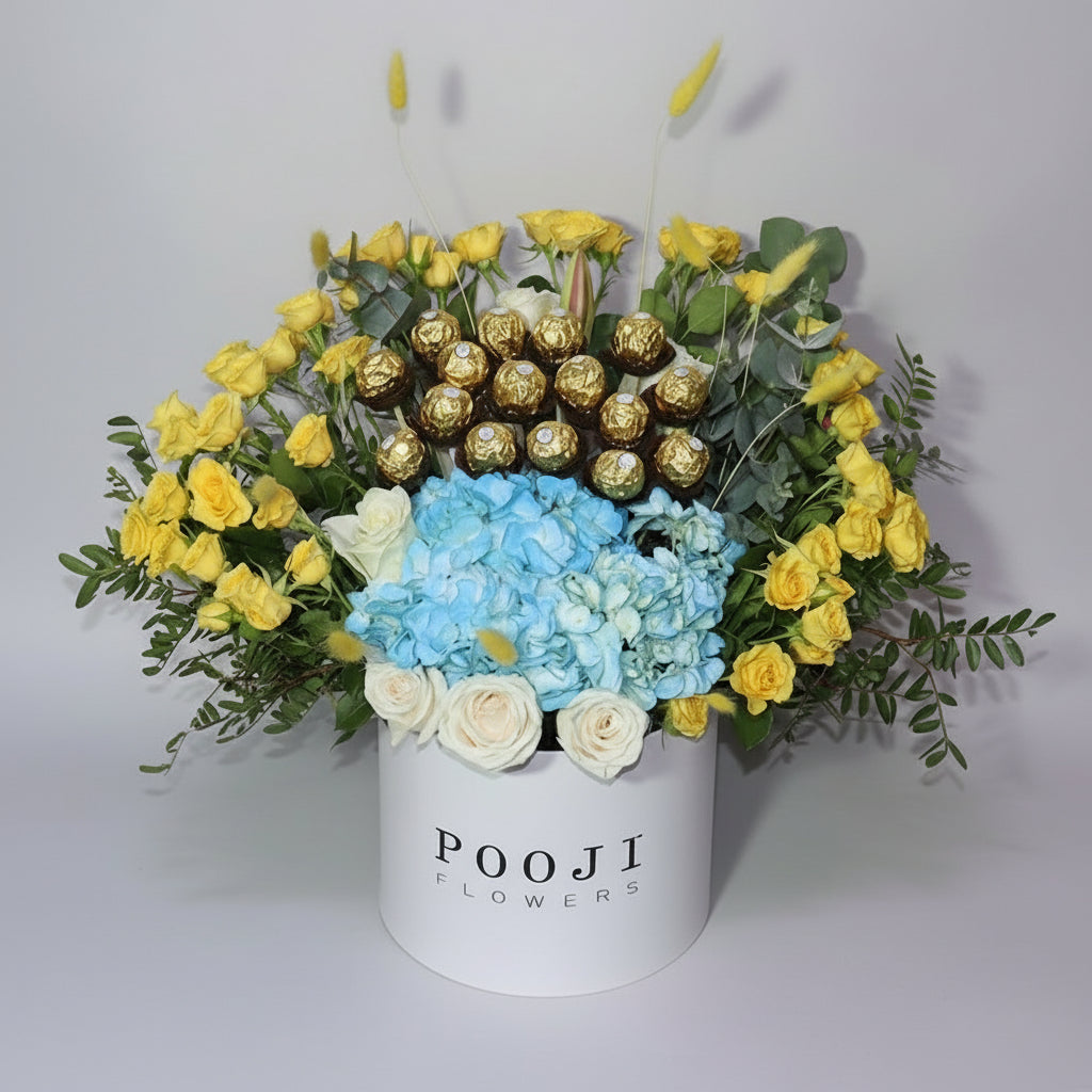Floral arrangement with yellow roses, blue hydrangeas, and gold chocolate bars in a white container labeled 'POOJI FLOWERS'.