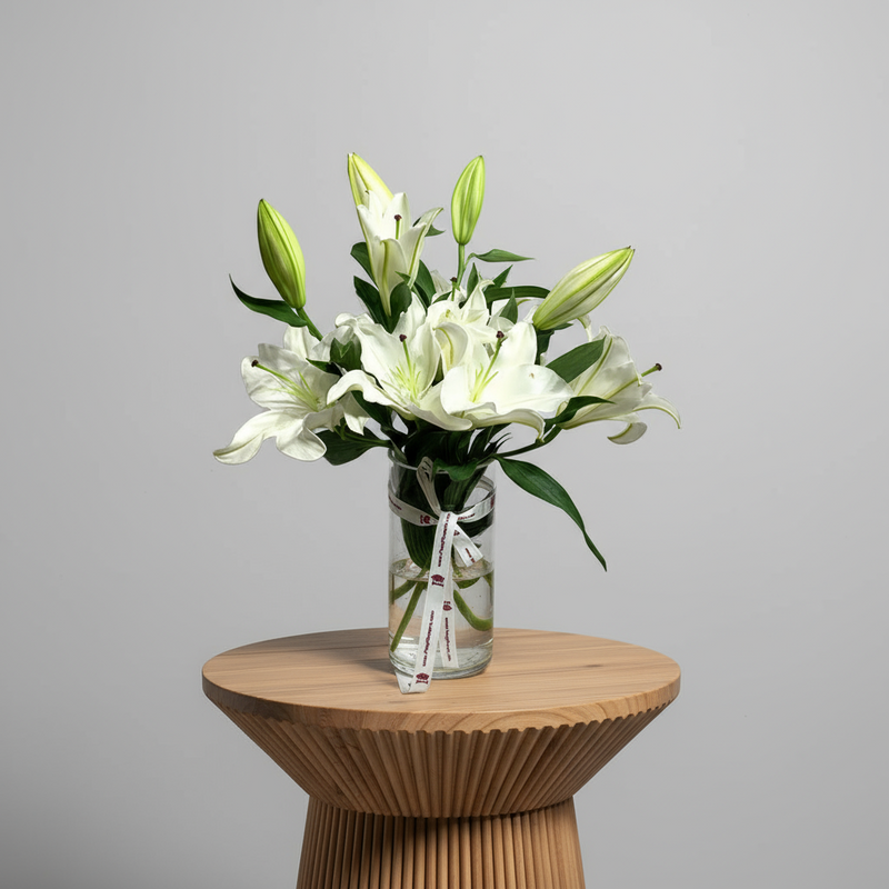 Elegant white lilies bouquet in a glass vase with green leaves and decorative ribbon