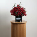 A bouquet of red flowers with green leaves, arranged in a black box with a label that says 'Pooji'.