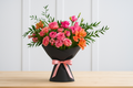 Bouquet of pink and orange flowers in a black vase with a pink ribbon on a wooden table.